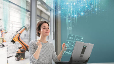 一个女人微笑着看着她的电脑，双手举在空中。笔记本电脑屏幕上会出现数字和图表。背景中可以看到一个制造机械臂