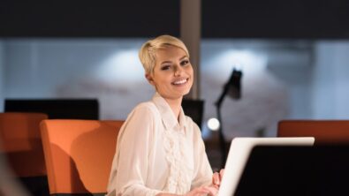 一个商务女性，在黑暗的办公室里，坐在办公桌前，对着笔记本电脑微笑